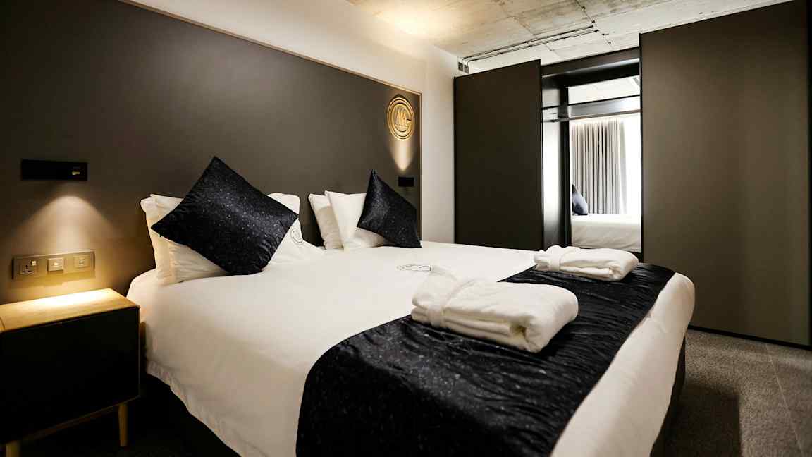 Sovrum med en modern dubbelsäng, vita sängkläder och mörka dekorativa kuddar. En nattduksbord med mjuk belysning står bredvid. (AI-genererad text)