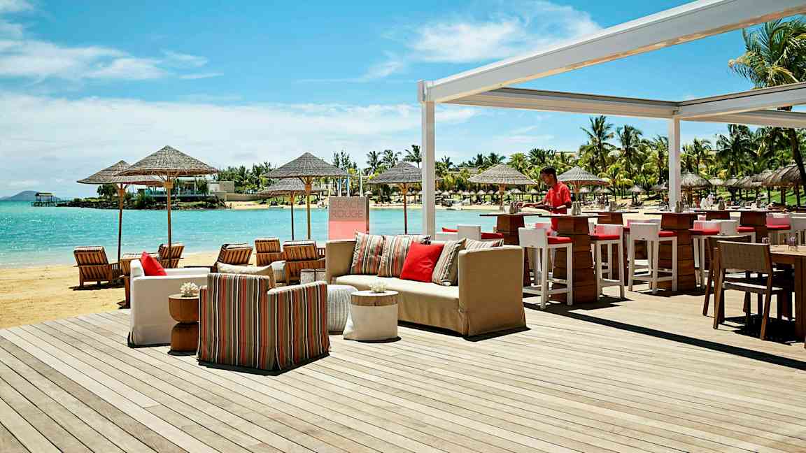 Uteservering vid stranden med bar och sittgrupper, samt utsikt över havet och omgivande palmer. (AI-genererad text)