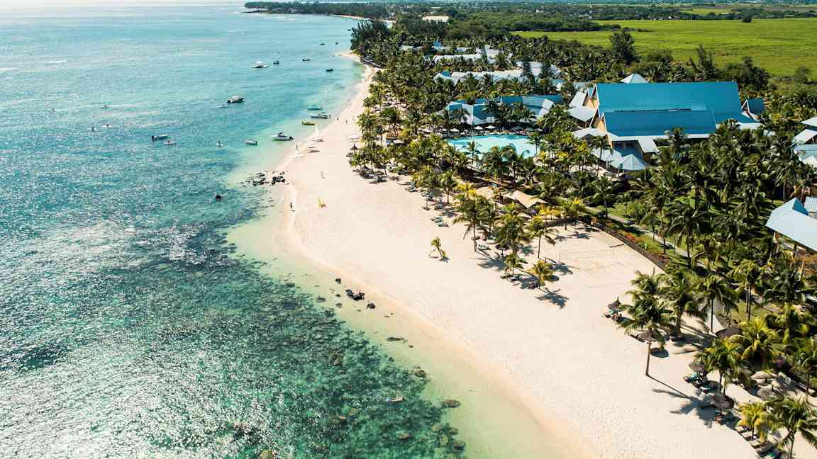 Utsikt över en kust med en bred sandstrand, turkost hav och ett poolområde omgiven av palmer och hotellbyggnader. (AI-genererad text)