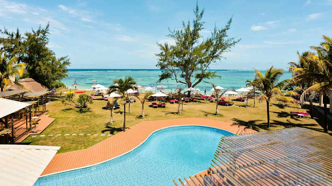 En pool med turkost vatten framför en gräsmatta med träd, solsängar och parasoller längs en strand. (AI-genererad text)