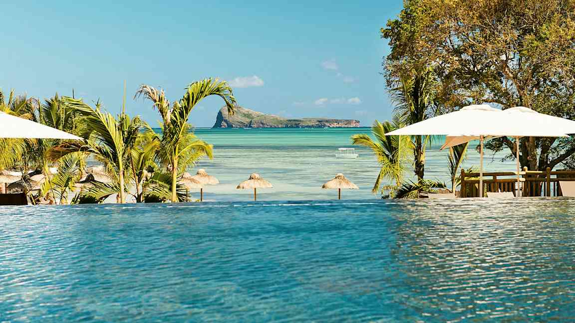 En infinitypool med utsikt över en strand med parasoller och en liten ö i havet. (AI-genererad text)
