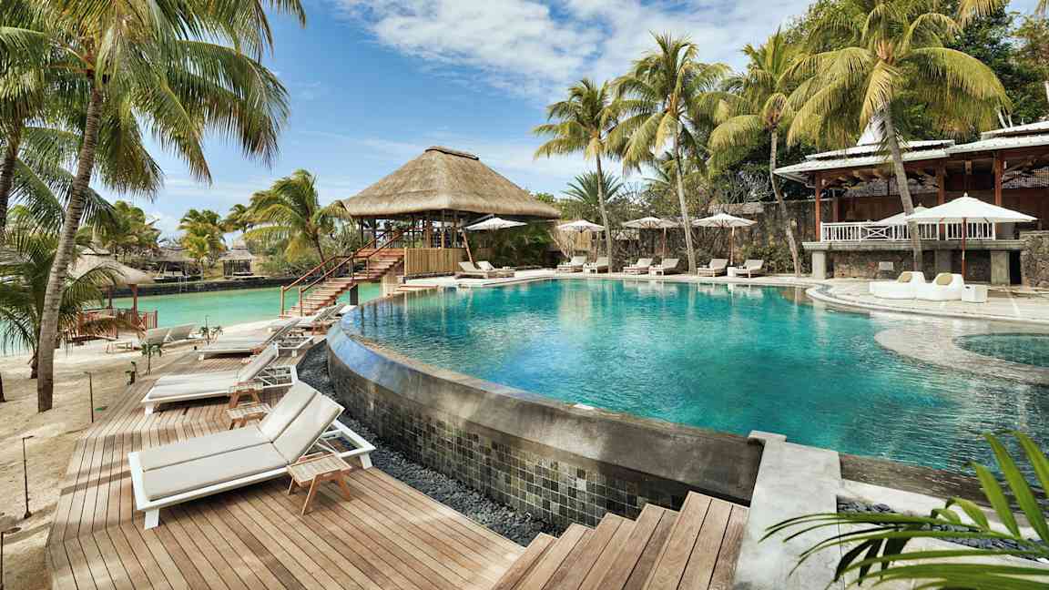 En pool omgiven av solsängar och parasoller, med palmer och en byggnad med halmtak i bakgrunden. (AI-genererad text)