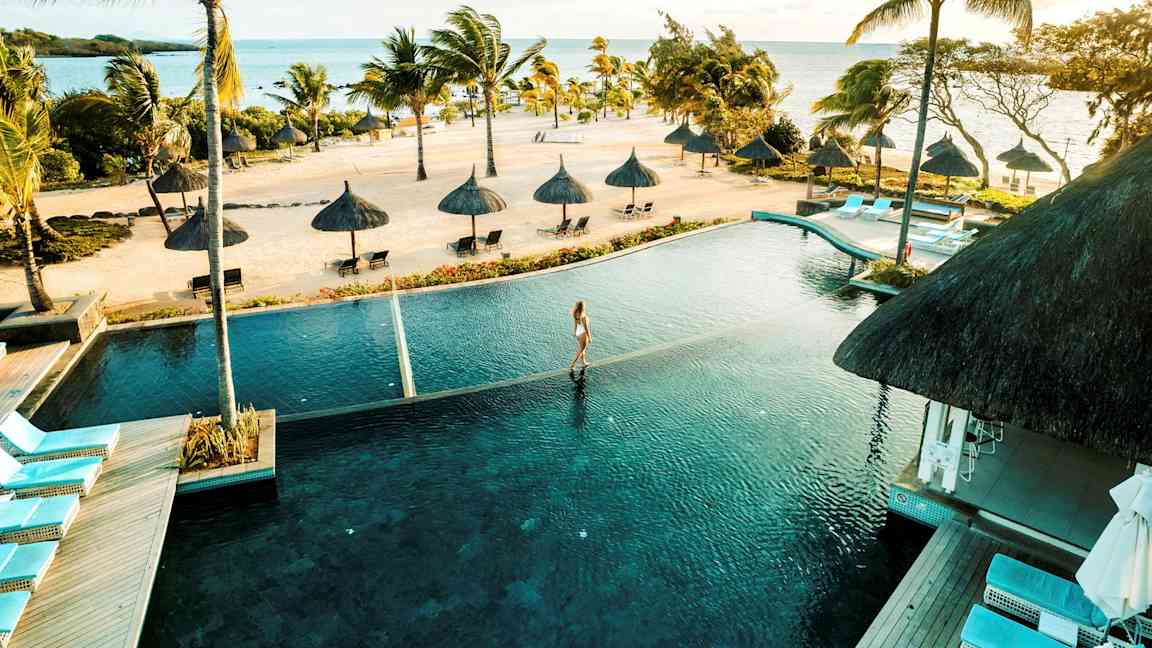 En tropisk strand med palmer och parasoller vid havet, en infinitypool i förgrunden. (AI-genererad text)