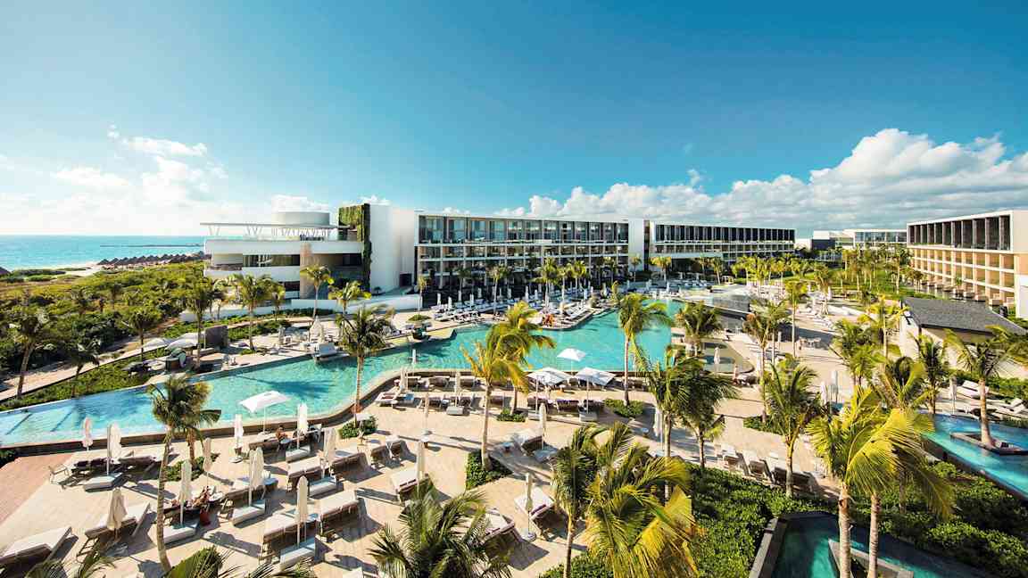 Ett modernt hotellkomplex med flera pooler, frodig vegetation och palmer, beläget vid kusten med utsikt över havet under en klarblå himmel. (AI-genererad text)