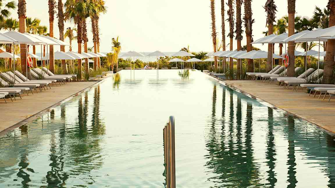 En avlång simbassäng omgiven av solstolar med parasoller och palmer. (AI-genererad text)