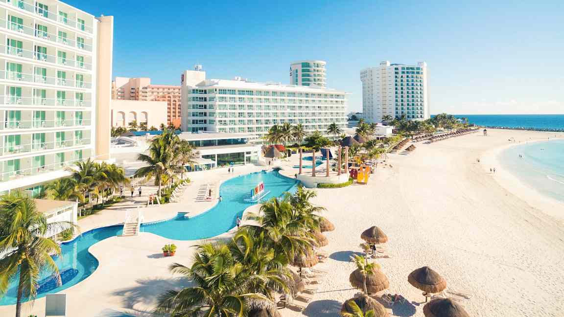Ett hotellkomplex vid en strand med pooler i förgrunden och havsvy i bakgrunden. (AI-genererad text)
