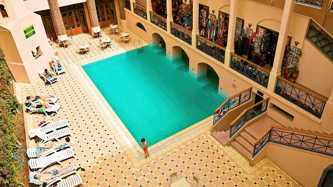 Ett inomhusområde med en swimmingpool omgiven av solstolar och bord under en balkong med affärer. (AI-genererad text)