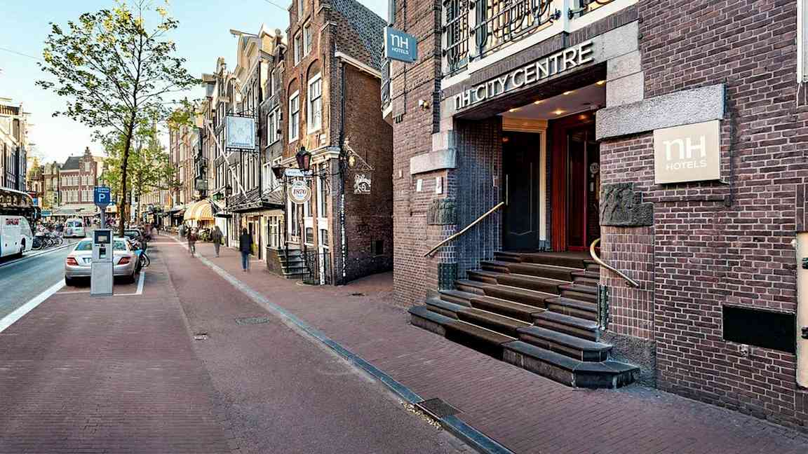 En gata med parkerade bilar och gångväg, omgärdad av byggnader i traditionell arkitektur där hotell NH City Centre har en synlig entré. (AI-genererad text)
