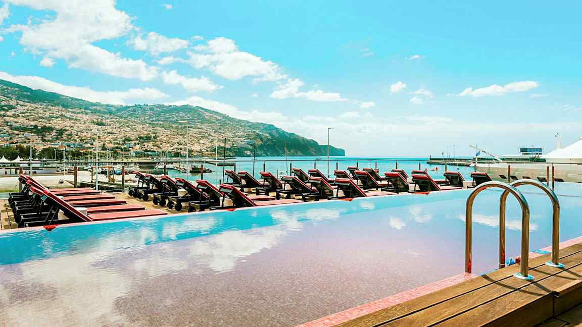 Infinitypool med utsikt över solstolar, havet, och en omgivande bergslandskap. (AI-genererad text)