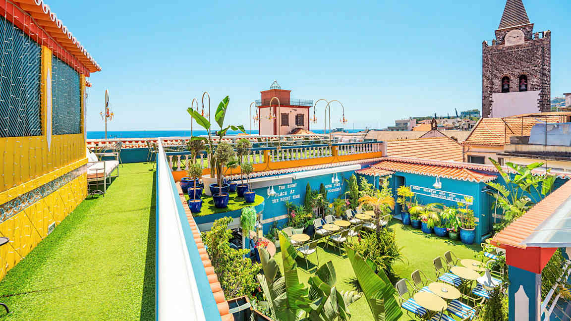 En takterrass med gröna växter, sittplatser och färgglada byggnader i bakgrunden. (AI-genererad text)