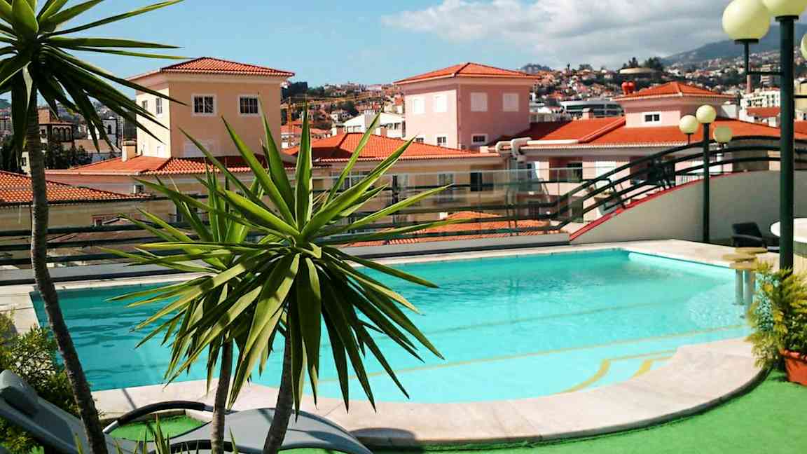 En pool omgiven av grönska och en bakgrund med hus med röda tegeltak. (AI-genererad text)