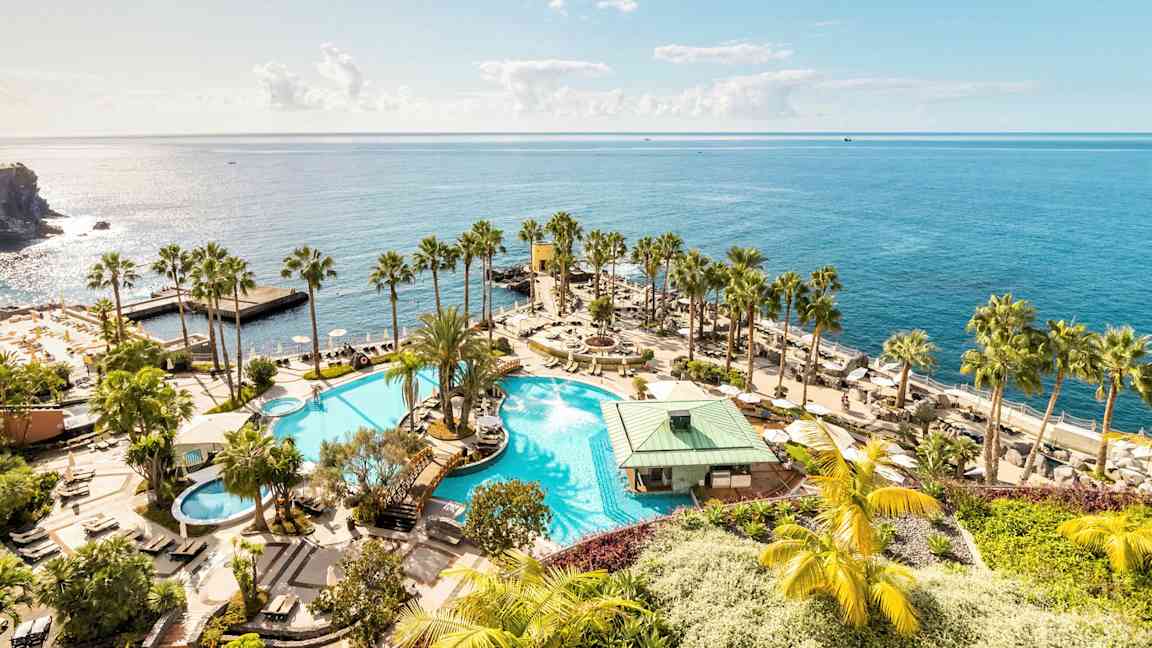 En stor pool med omgivande palmer och solstolar, belägen vid stranden med utsikt över det glittrande havet. (AI-genererad text)