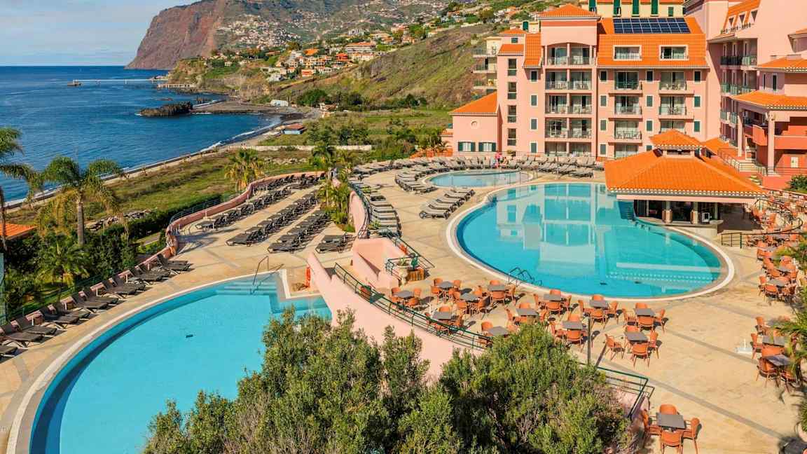 Ett hotellområde vid kusten med två stora pooler, solstolar, bord, och omgivande byggnader med orange tak. Bakgrunden visar en klippa vid havet och bostadsområden längs sluttningen. (AI-genererad text)