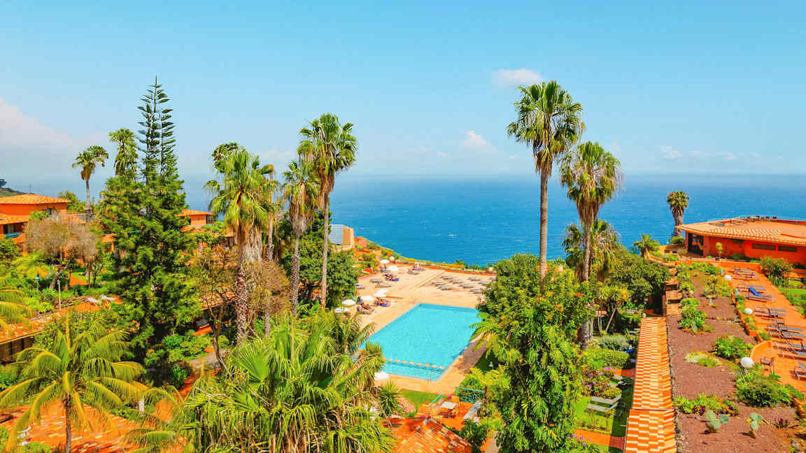 Utsikt över ett poolområde omgivet av grönska och palmer, med havet i bakgrunden. (AI-genererad text)