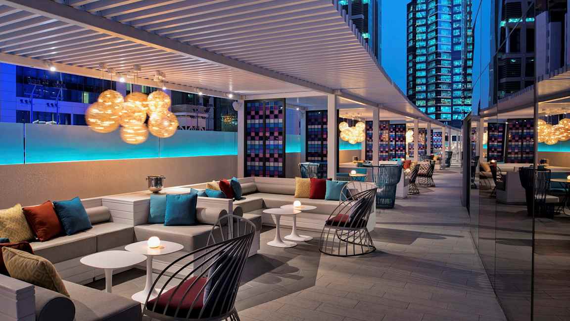 En modern lounge med sittgrupper, soffor och bord, dekorerad med färgglada kuddar och lysande lampor, omgiven av höga byggnader på natten. (AI-genererad text)