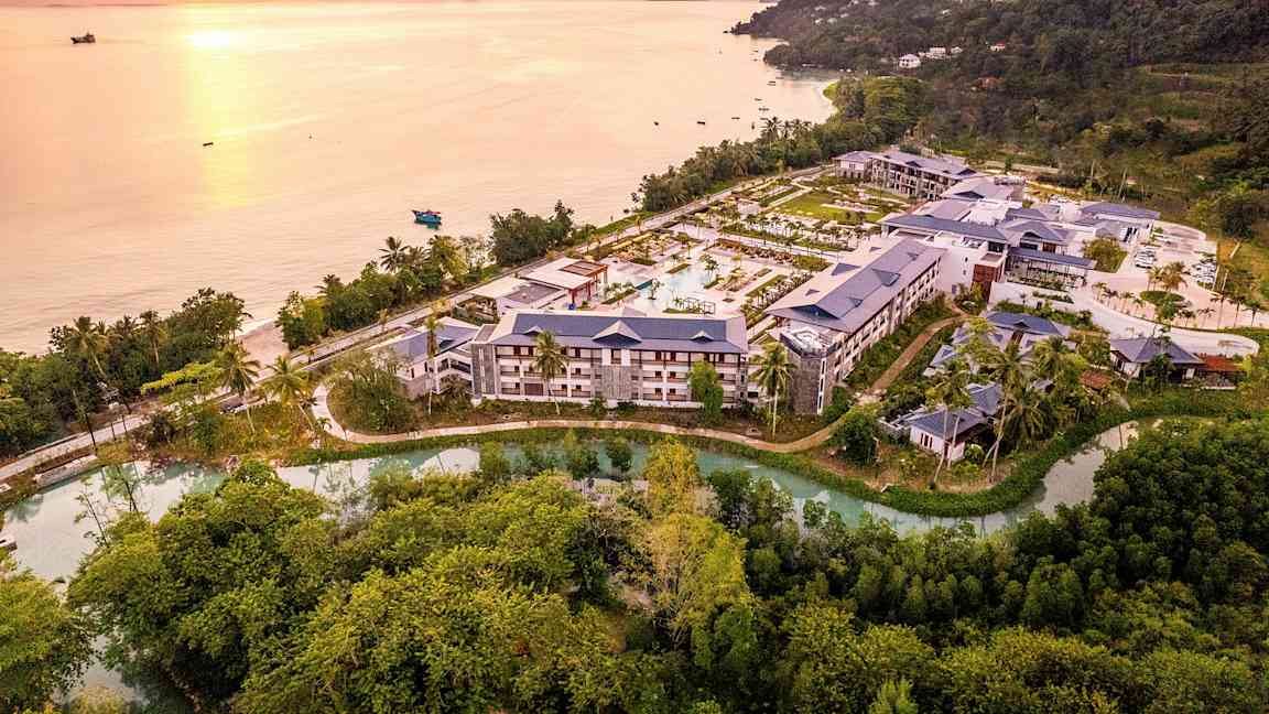 Flygbild över ett hotellkomplex omgivet av natur och beläget nära en kust med strand och hav vid solnedgång. (AI-genererad text)