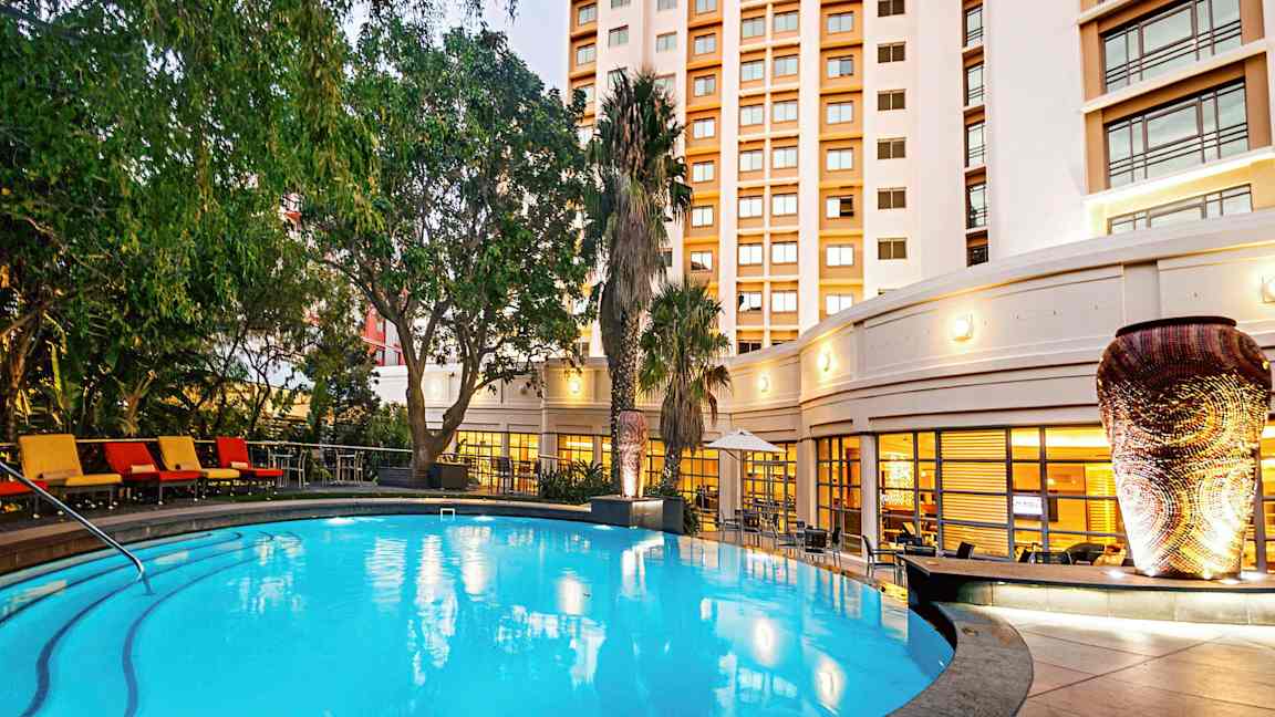 En upplyst utomhuspool omgiven av gröna växter och en byggnad i bakgrunden. (AI-genererad text)