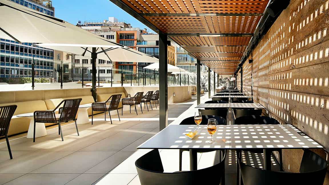 En utomhusterrass med bord och stolar under ett träpergola och stora parasoller. (AI-genererad text)