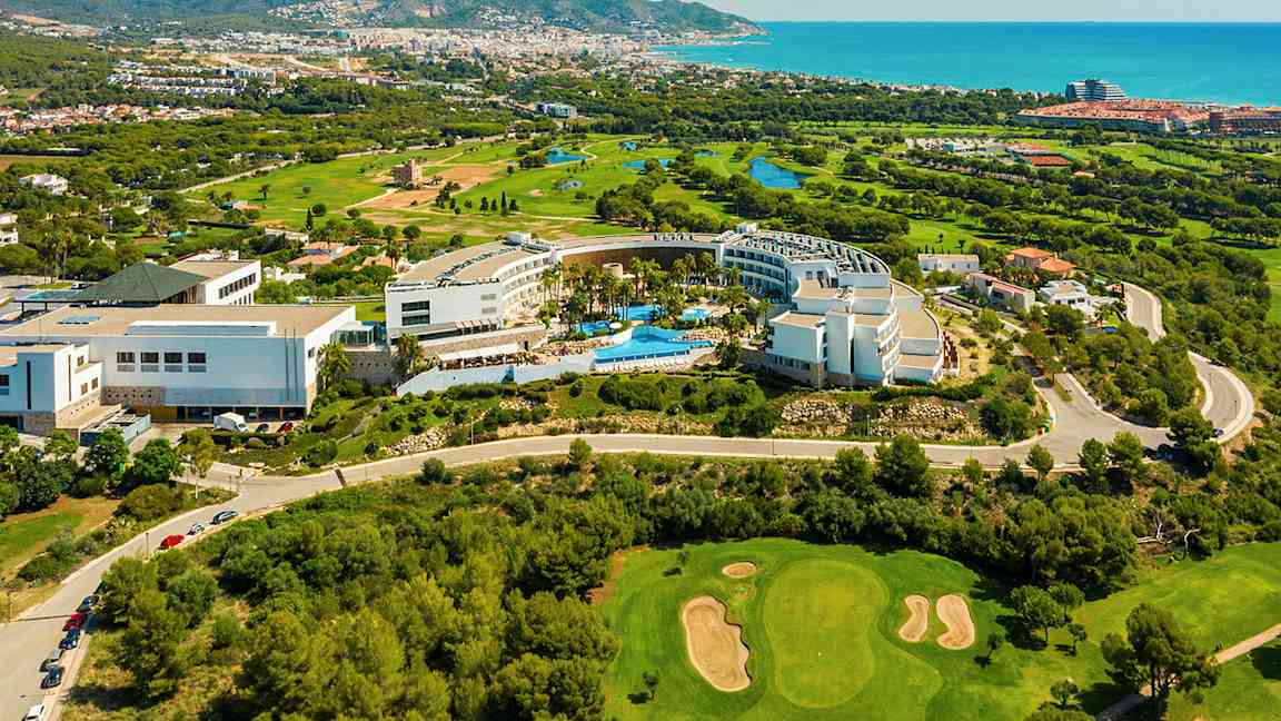 Vy från ovan av ett hotellkomplex omgivet av grönska och en golfbana. Havet och en stad ligger i bakgrunden. (AI-genererad text)