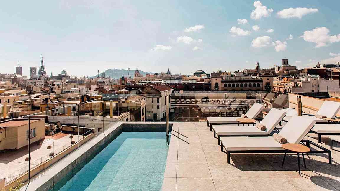 En utomhuspool och solstolar på en takterrass med utsikt över en stad och dess byggnader, under en klar himmel. (AI-genererad text)