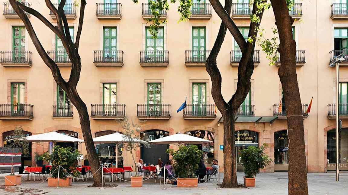 Ett hotell med flera våningar och balkonger, en trottoar med parasoller och sittplatser bland träd. (AI-genererad text)