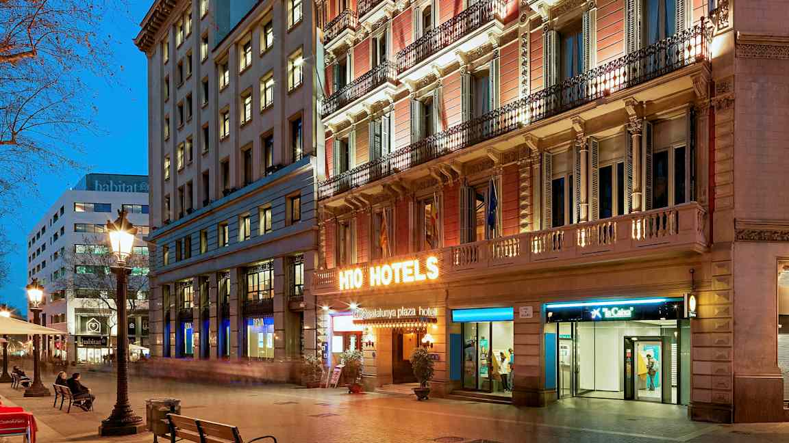 En stadsgata vid skymning med upplysta byggnader. En hotellskylt lyser starkt på fasaden. (AI-genererad text)