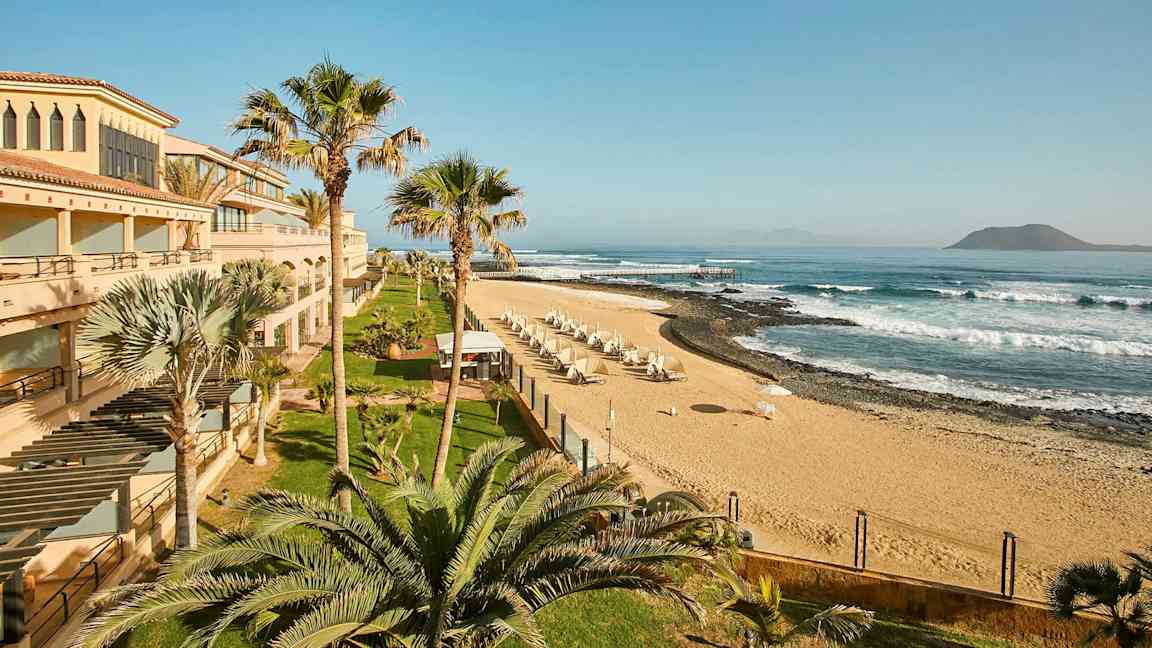 En strand med sand, parasoller och vågor, omgiven av byggnader och palmer. (AI-genererad text)