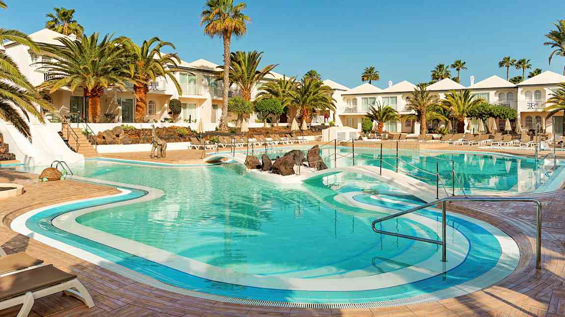 En hotellpool omgiven av palmer, solstolar och vita byggnader. (AI-genererad text)