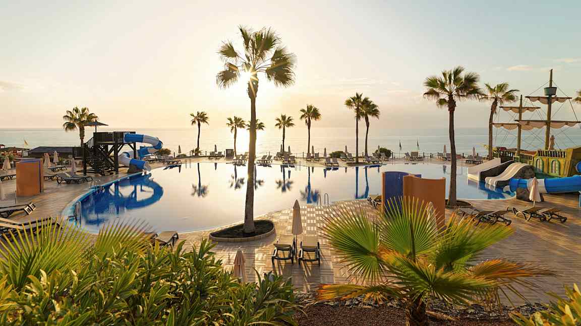 Bild på en pool med vattenrutschbanor, solstolar och palmer, samt havet i bakgrunden vid solnedgång. (AI-genererad text)