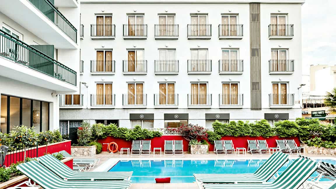 Bild på en hotellbyggnad med vit fasad och flera balkonger. Framför byggnaden finns en utomhuspool med randiga solstolar placerade runt. (AI-genererad text)