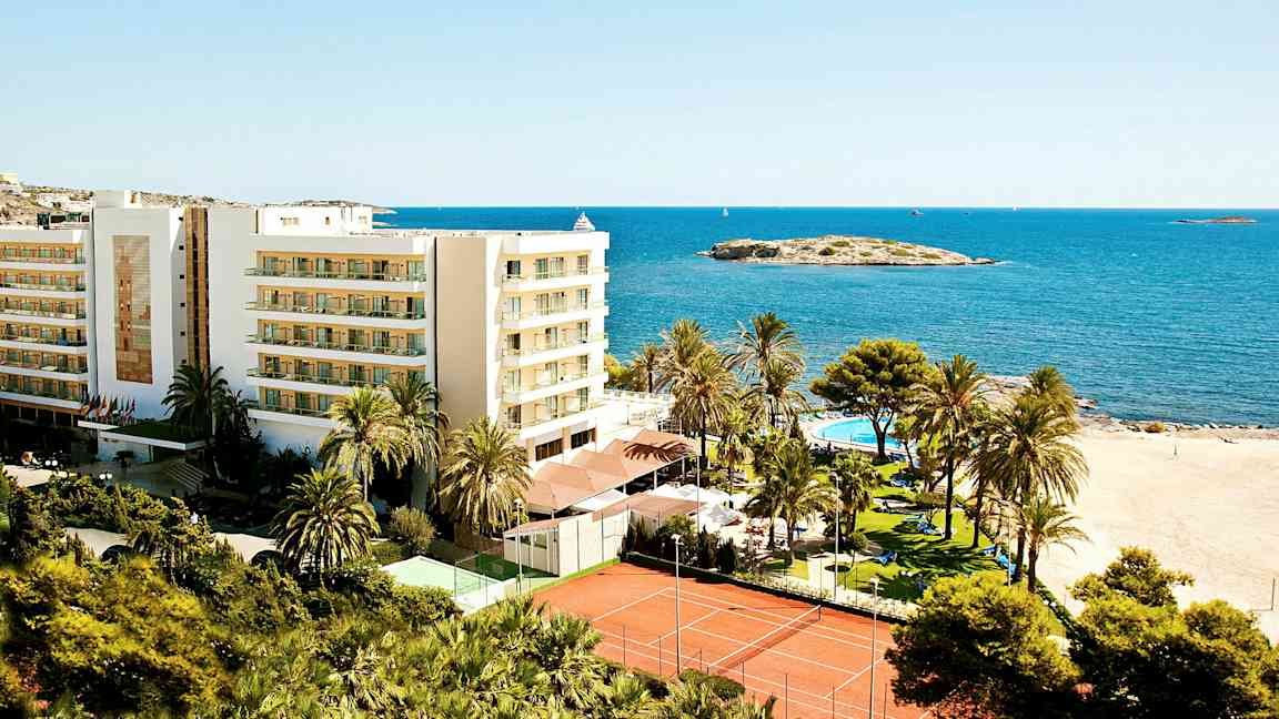 Bild av en semesteranläggning intill stranden med en byggnad, palmträd, pool och tennisbanor, belägen vid havet. (AI-genererad text)