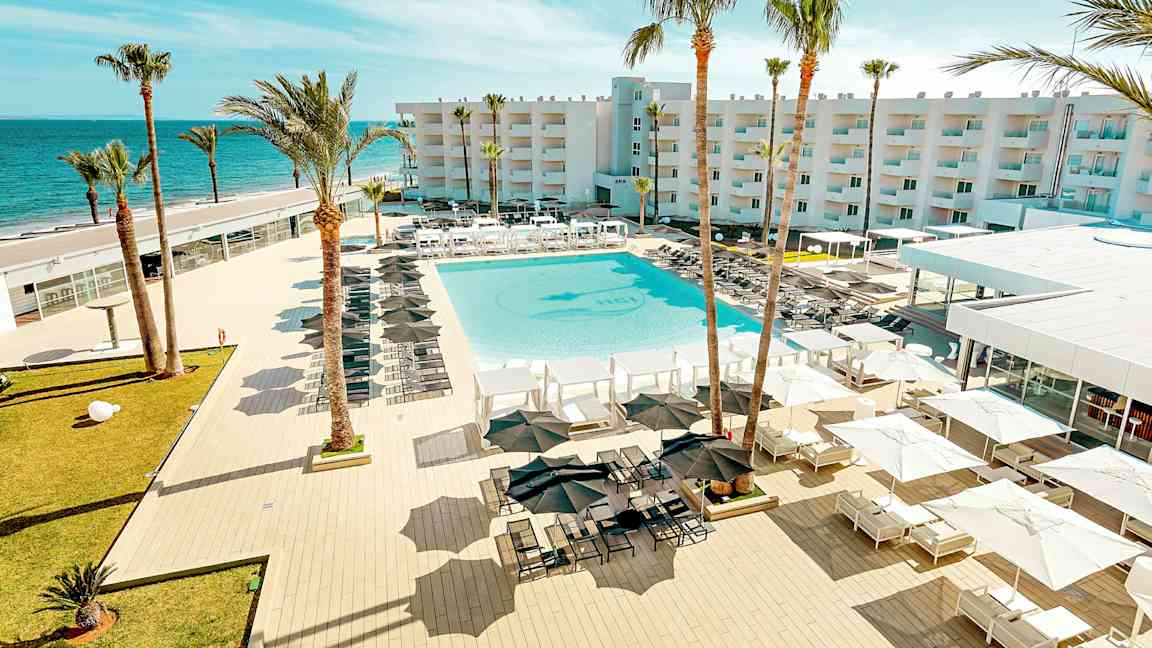 En vy över en resort med en stor pool, omgiven av solstolar och parasoller, med palmer och en byggnad i bakgrunden nära havet. (AI-genererad text)