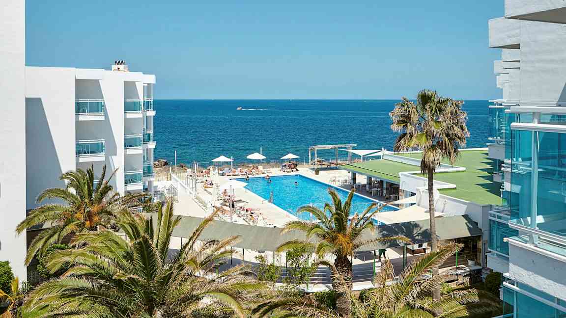 En bild av ett hotellområde med utsikt över havet. Det finns en swimmingpool omringad av solstolar och parasoller, omgiven av palmer och vita byggnader. (AI-genererad text)