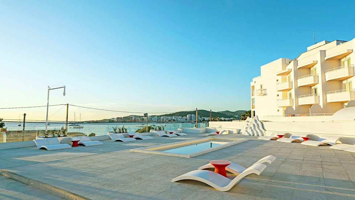 Utomhuspoolområde med moderna solstolar och utsikt mot havet samt vita byggnader i bakgrunden. (AI-genererad text)