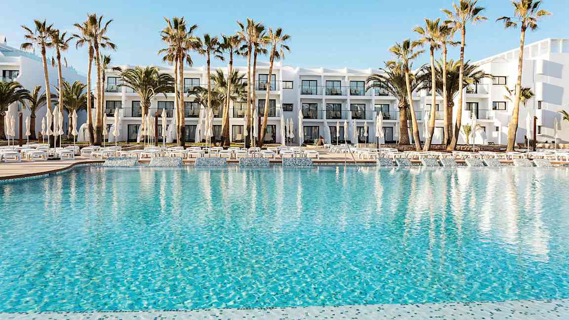 Hotellets poolområde med klarblått vatten, omgivet av solstolar, parasoller och palmträd. I bakgrunden syns en vit byggnad med balkonger. (AI-genererad text)