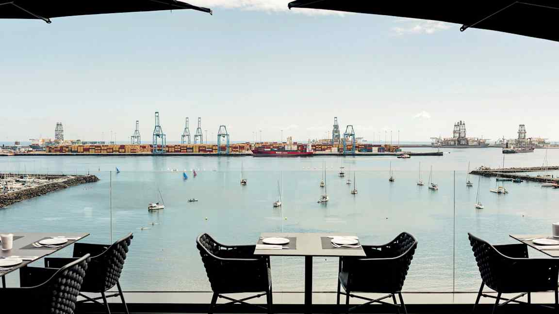 Utsikt över en hamn med kranar och förtöjda båtar. Förgrunden visar bord och stolar från ett café. (AI-genererad text)