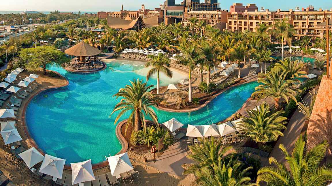 En stor utomhuspool omgiven av parasoller, solstolar och grönska vid ett semesterhotell. (AI-genererad text)