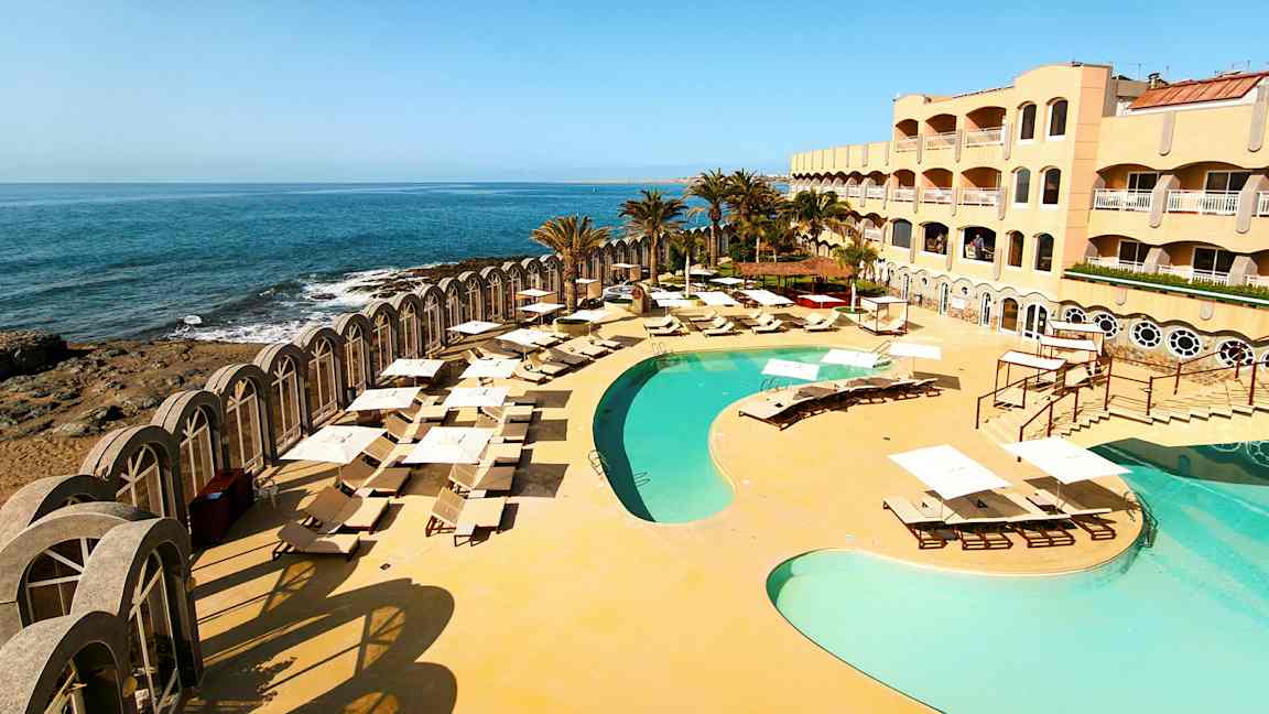Bild av ett hotellkomplex med pool och solstolar vid havet. (AI-genererad text)