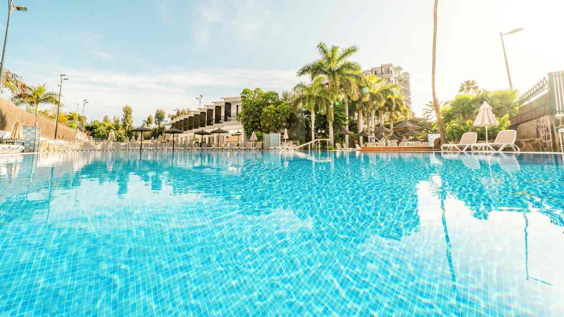 Bild av en utomhuspool omgiven av palmträd och solstolar. (AI-genererad text)