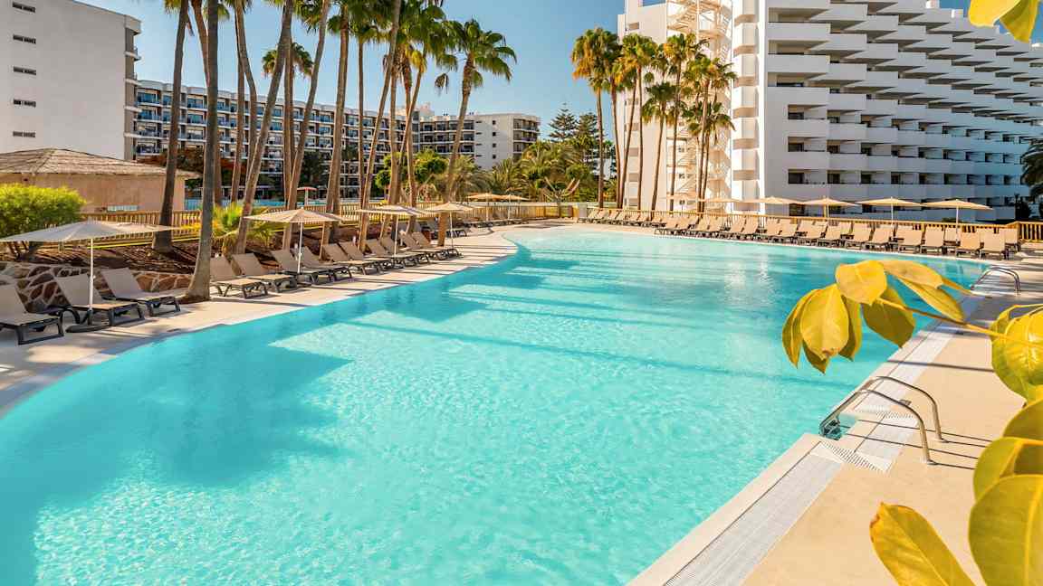 En stor swimmingpool omgiven av solstolar, parasoller och palmer vid ett hotell. (AI-genererad text)