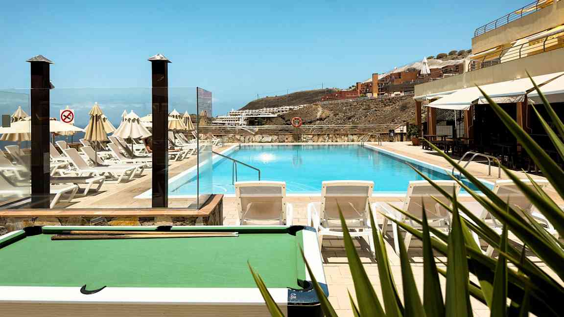 Utomhuspool med omgivande solstolar och parasoller, med utsikt över bakgrunden av byggnader på en sluttning. (AI-genererad text)