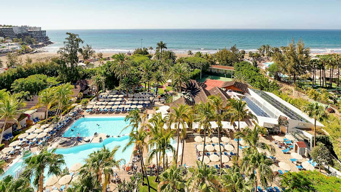 Utsikt över ett semesterkomplex med pooler, solstolar och palmer nära en sandstrand vid havet. (AI-genererad text)