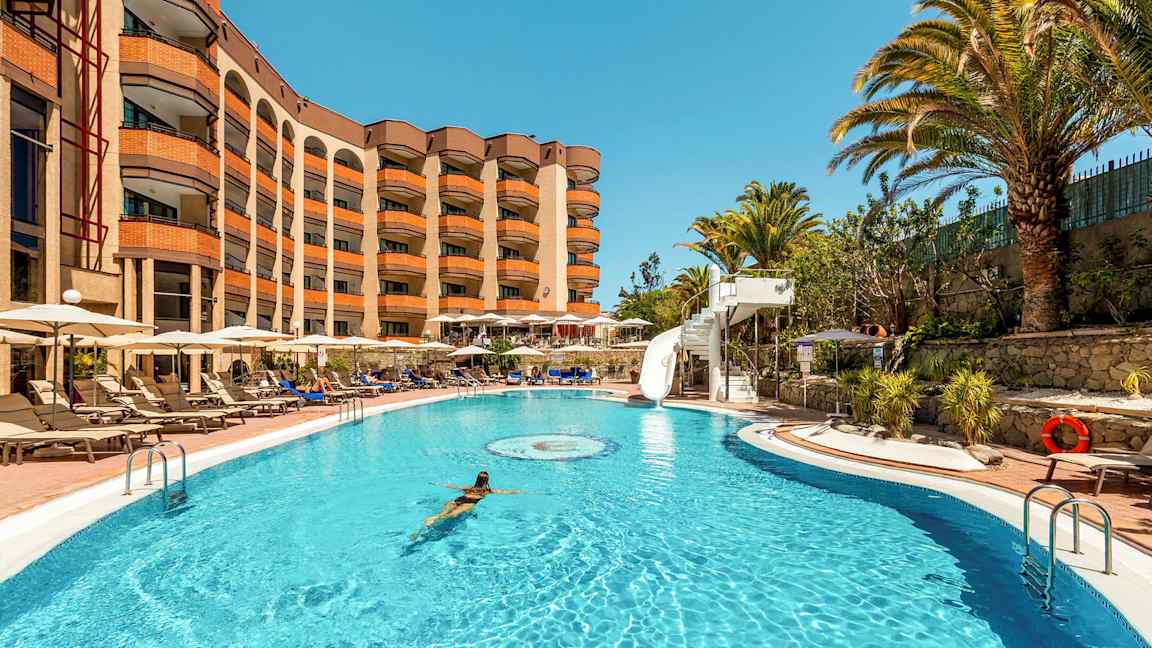 En pool med en kvinna som simmar, omgiven av solstolar och ett hotell i bakgrunden. (AI-genererad text)