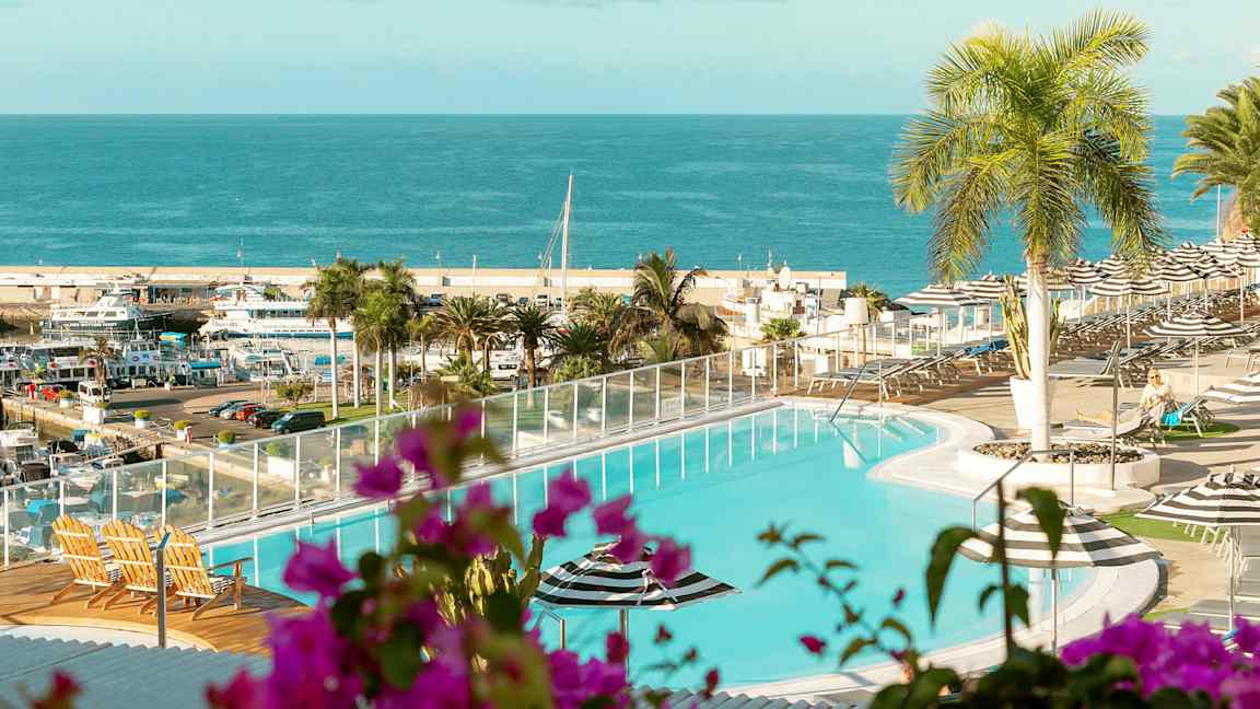 En pool med utsikt över ett hav och en hamn. I förgrunden finns växter och utemöbler. (AI-genererad text)