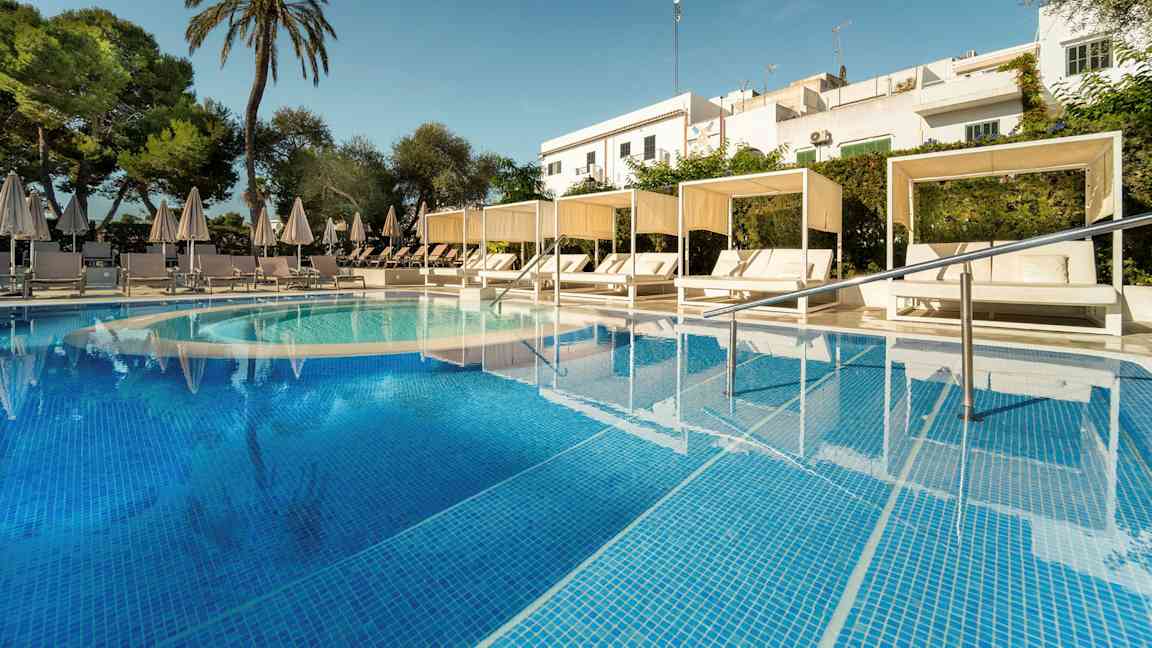 Bild av en pool med turkost vatten och vita solsängar under parasoll samt cabanas. Det är omgivande grönska och en skymt av byggnader i bakgrunden. (AI-genererad text)