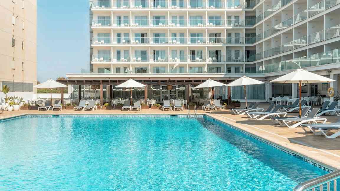 En byggnad med flera balkonger, framför en pool med solstolar och parasoller. (AI-genererad text)