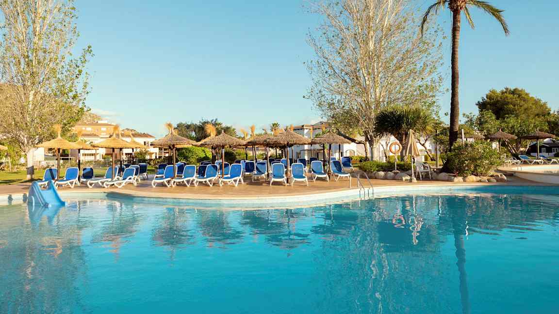 En pool vid ett hotell omgiven av solstolar och parasoller, omgärdad av träd och grönska. (AI-genererad text)