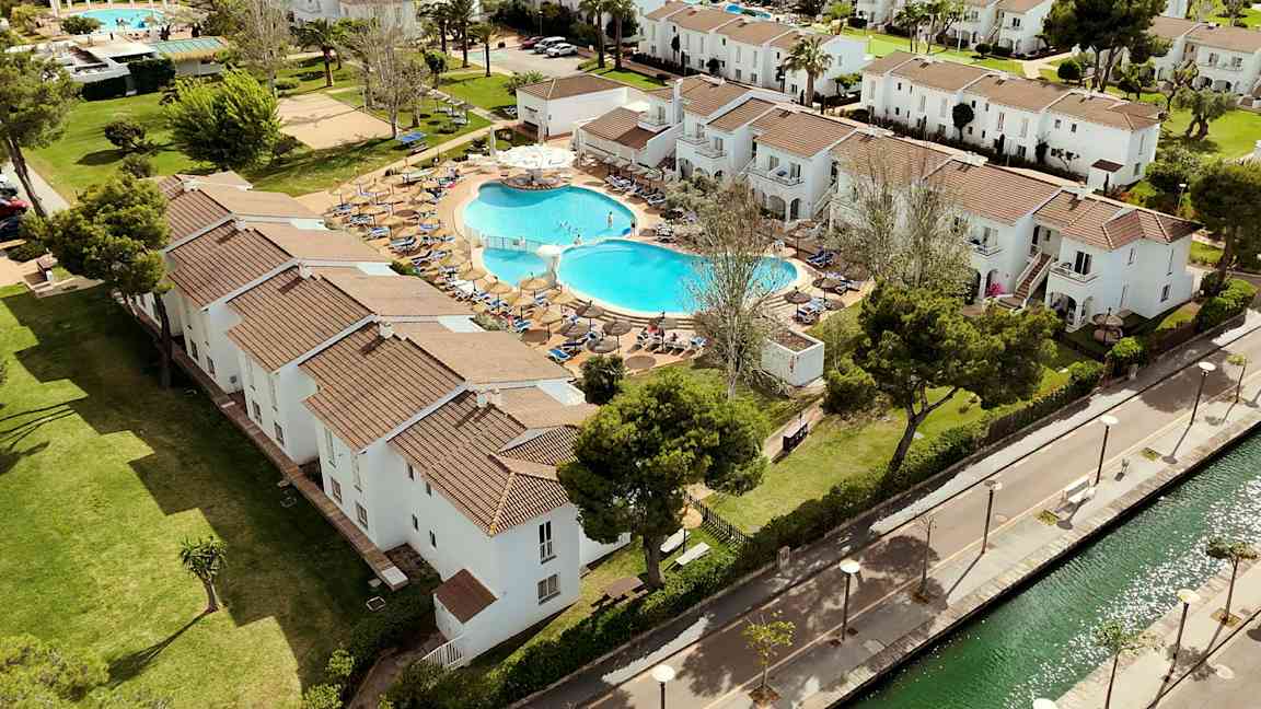 En flygbild av ett semesterkomplex med vita byggnader, en pool och grönområden. (AI-genererad text)