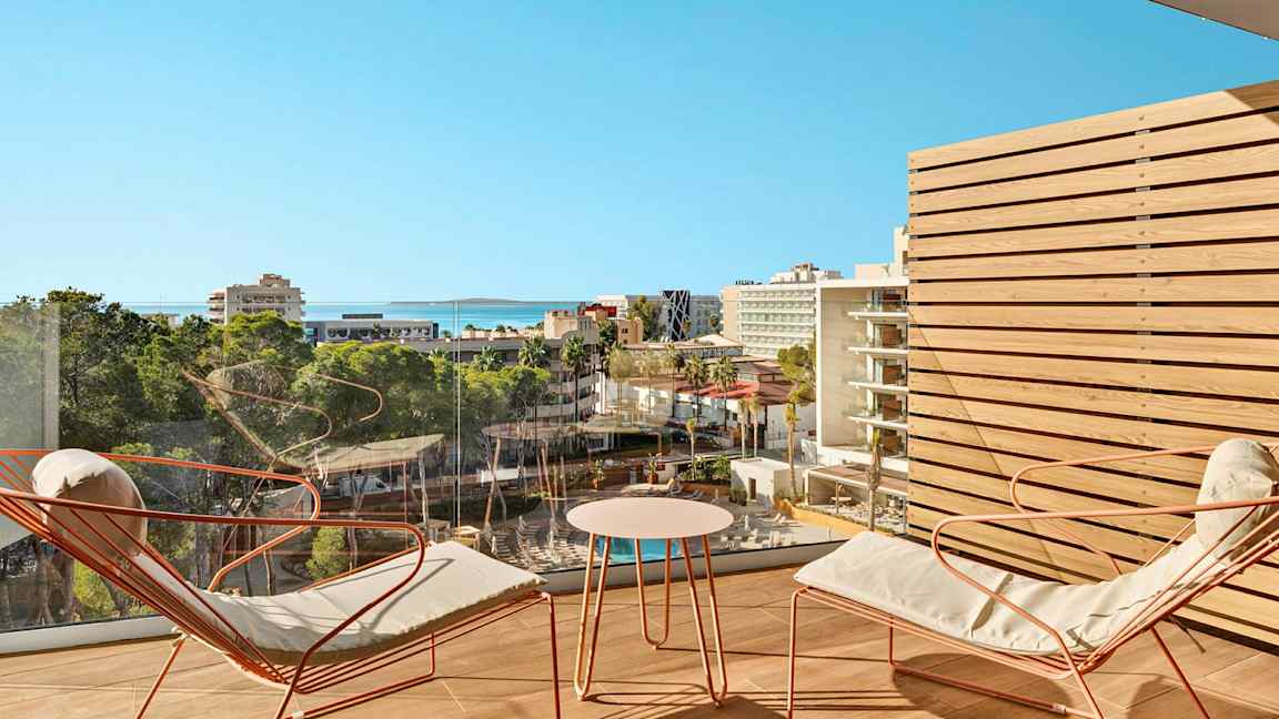 Vy över ett stadsmiljö med hotell och grönområden från en balkong med sittmöbler. (AI-genererad text)