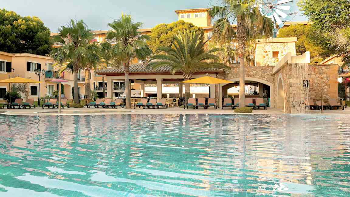 En pool framför en byggnad med palmer och en liten vindmölla synliga. (AI-genererad text)